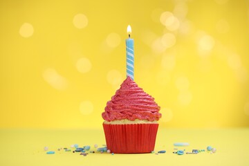 Burning candle in cupcake and sprinkles on yellow background with blurred lights