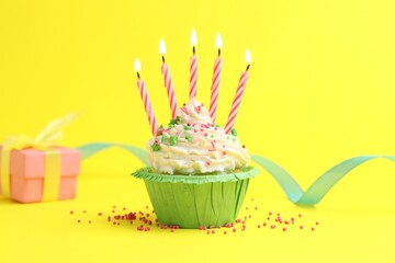 Burning candles in cupcake, sprinkles and party decor on yellow background