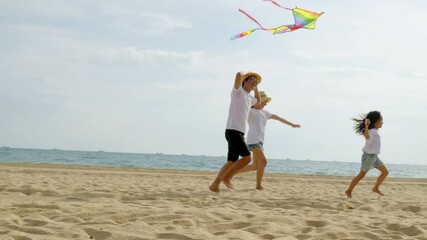 Asian family is flying a kite high in the sky while running on the beach. Happy parents mother and father with their child playing with kite, family is enjoying their time together, summer day