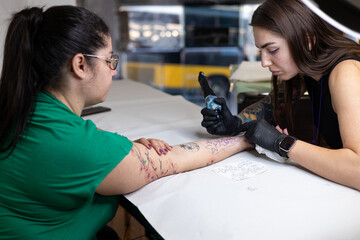 Close-up of a tattoo artist creating a floral tattoo on a woman's hand using a tattoo machine in...