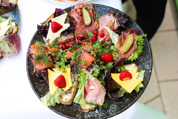 Gourmet open sandwiches with assorted meats, cheeses, smoked salmon, pickles, lettuce, and strawberries served on a dark rustic plate at a catered event.