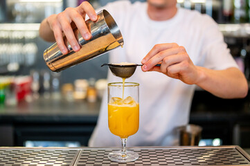 Close-up of a bartender straining a vibrant orange cocktail into a glass with ice, set against a...