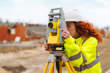 Young female surveyor operates a total station at a construction site while wearing safety hi-viz jacket