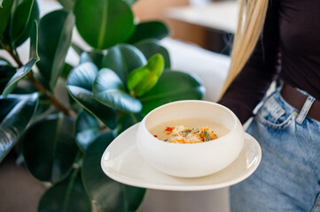 bowl of creamy soup garnished with herbs, held by hands over a white plate, against a green leafy background. Minimalist food presentation, natural light.