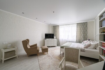 cozy bedroom with a neutral palette, featuring a bed, armchair, TV, and natural light streaming through the window. A calm and inviting space