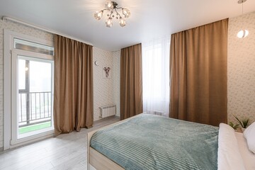 Cozy bedroom with light-colored walls, warm brown curtains, a blue-striped bedspread, and a balcony door