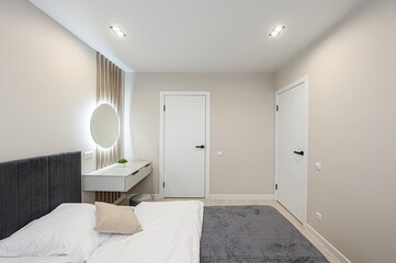 Cozy bedroom featuring a bed with a gray headboard, illuminated round mirror above a white vanity, and two modern white doors