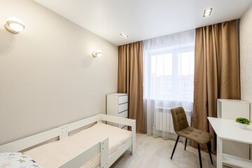 cozy room featuring a white wooden bed frame, a desk, a chair, and a window with sheer and brown curtains. Wall sconces provide soft lighting in the neutral-toned space