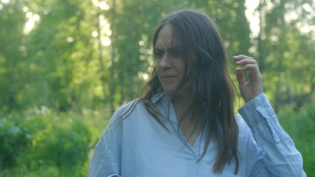Urban woman waist-up in grassy forest endures mosquito swarm attacking bare arms and neck. Frantically swiping at insects while face shows discomfort from relentless summer bites in wilderness. 