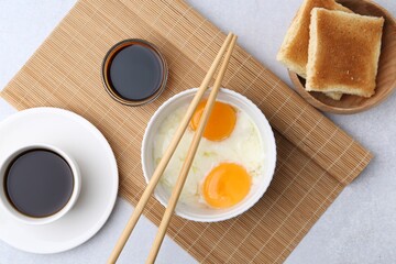 Half-boiled eggs in bowl, soy sauce, chopsticks, coffee and toasted bread on light grey table, flat lay