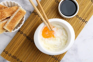 Half-boiled egg in bowl, soy sauce, chopsticks and toasted bread on white marble table, flat lay