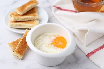 Half-boiled egg in bowl and toasted bread on white marble table, closeup