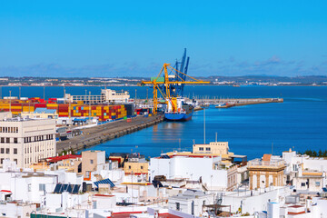 Fototapeta premium Port of Cadiz is bustling with activity, colorful shipping containers, towering cranes, and large cargo ship docked at pier. Rooftops of buildings line foreground, calm waters extend toward horizon
