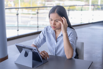 Anxious Asian woman experiencing a headache or stress while working on her tablet, capturing the challenges of remote work and mental strain in a contemporary setting