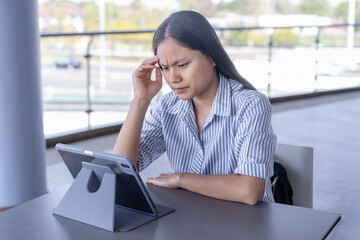 Anxious Asian woman experiencing a headache or stress while working on her tablet, capturing the challenges of remote work and mental strain in a contemporary setting