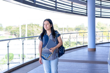 Thoughtful Asian woman standing with a tablet and bag, looking out into a modern, bright open space. Perfect for themes of contemplation, professional lifestyle, and urban environments.