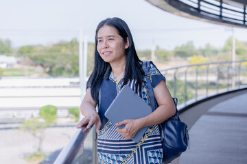 Thoughtful Asian woman standing with a tablet and bag, looking out into a modern, bright open...