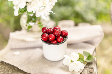 Enamel mug with cherries and white jasmine flowers in vase on towel-covered surface. Soft morning...
