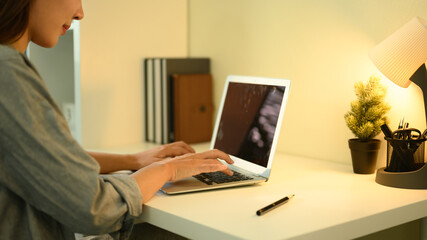 Close-up of a woman typing on a laptop at a home office desk with warm lighting