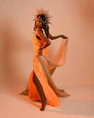 Full length portrait of a female model wearing a  fantasy crown, yellow gown and flowing fabric cape.  Walking pose on studio background with warm golden lighting