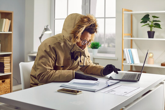 Young African American man freezing in cold office. Warmly dressed focused businessman wearing winter jacket and gloves analysing financial documents at workplace. Heating problems, power crisis