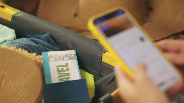 Traveler booking hotel online using smartphone sit at home beside packed suitcase, passport, ticket, clothes, straw hat displayed.Traveling and vacation concept 