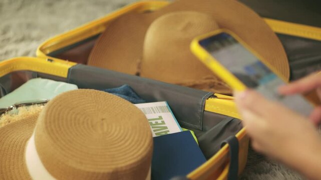 Traveler booking hotel online using smartphone sit at home beside packed suitcase, passport, ticket, clothes, straw hat displayed.Traveling and vacation concept