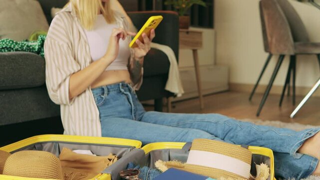Woman sitting on floor near open suitcase, browsing smartphone and organizing summer travel essentials with relaxed, anticipatory mood while preparing for upcoming trip.Traveling and vacation concept