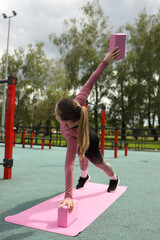 A young woman performs a yoga pose in a park, utilizing pink blocks for support. She balances...