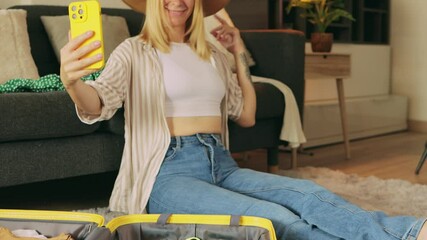 Blond traveler adjusting summer hat, packing luggage and taking selfies before vacation departure, smartphone in hand.Travel, blogging, weekend, relax and lifestyle concept