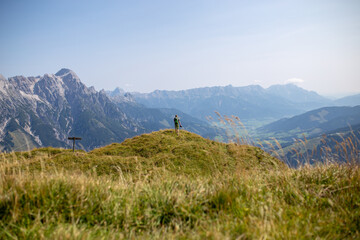 A hiker with a backpack and camera enjoys the breathtaking view from a grassy peak, with majestic mountains on the horizon.