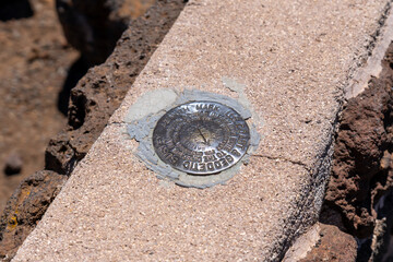 Geodetic survey marker embedded in concrete, surrounded by dark rocks, indicating a precise...