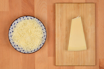 A top-down view displays a bowl of grated cheese beside a triangular wedge of cheese on a wooden...