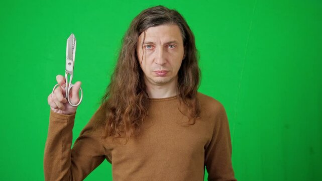 A serious and direct mood is captured as a man with long hair holds large tailor's shears, looking directly at the camera against a vibrant green screen. It's ideal for themes of craftsmanship