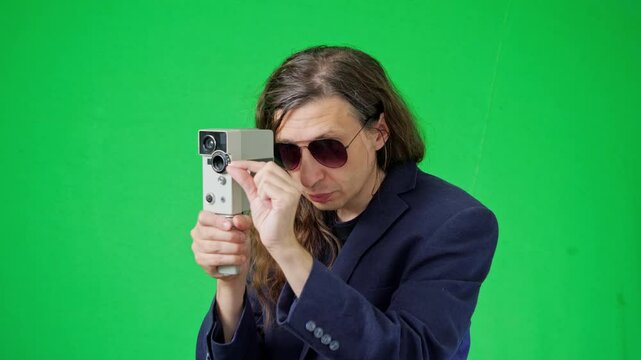 A focused and artistic mood is captured as a man in sunglasses and a suit jacket meticulously adjusts a vintage film camera, looking down at its controls against a vibrant green screen