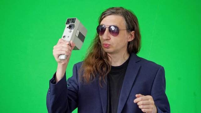 A focused and artistic mood is captured as a man in sunglasses and a suit jacket meticulously adjusts a vintage film camera, looking down at its controls against a vibrant green screen