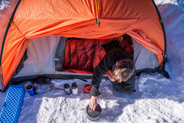 Man lying in sleeping bag cooks on portable stove outside winter tent in snowy campsite.Top-down view of cold weather camping gear © XArt
