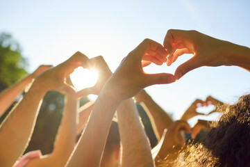 Group of people making heart shape gesture standing in summer park at sunset. Thankful young friends doing love sign raising hands up in nature against blue sky. Friendship, care and people concept