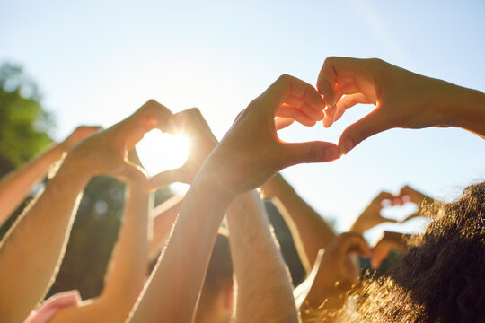 Fototapeta Group of people making heart shape gesture standing in summer park at sunset. Thankful young friends doing love sign raising hands up in nature against blue sky. Friendship, care and people concept