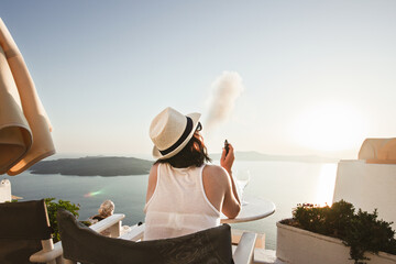 A woman enjoys a scenic sunset view over the Aegean Sea from a Santorini terrace with a glass of wine and a vape.