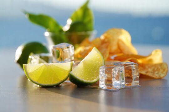 Close up of lime wedges ice cubes and potato chips on a reflective surface with a blurry background