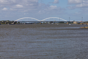 The Merwedebrug Gorinchem (Merwede Bridge) arches gracefully over a wide river, bustling with boats and traffic, under a bright, cloudy Dutch sky.