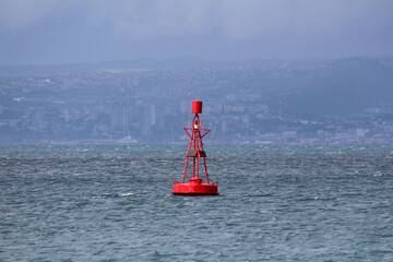 Bright red navigation buoy with solar panels and cage top floats on a choppy sea, distant city skyline visible through haze and low clouds across the water.