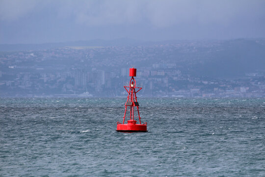 Bright red navigation buoy with solar panels and cage top floats on a choppy sea, distant city skyline visible through haze and low clouds across the water.