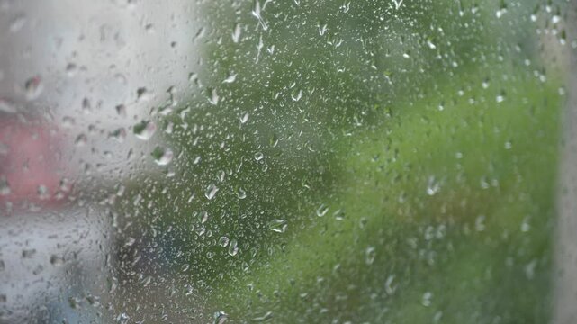 Close-up shot of raindrops falling on a window during a rainy day. The focus is on the water droplets running down the glass, with a soft, shallow depth of field.