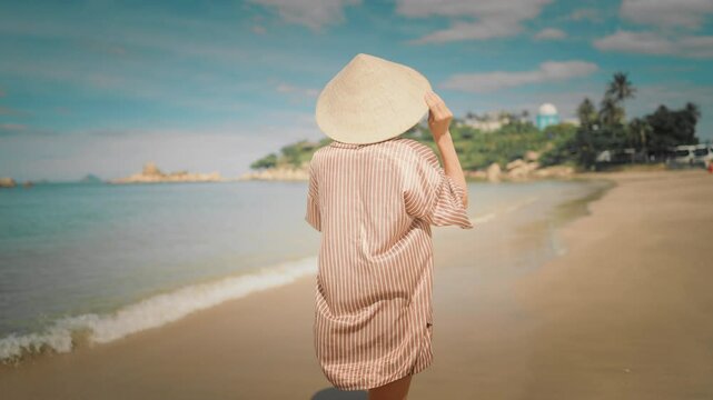 Summer trip to Vietnam back view of slender woman in traditional hat on beach. Medium shot with carefree female vacationer relaxing and walking on beautiful seacoast in sunny day, vacation trip