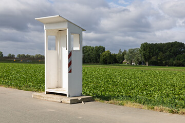 An empty white booth, a guard booth, stands by a rural road with a vibrant green field and cloudy sky in the background.
