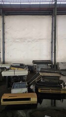 Broken air conditioning units are piled up inside a warehouse, resting on a damp concrete floor with water puddles and a stained, worn-out wall in the background.