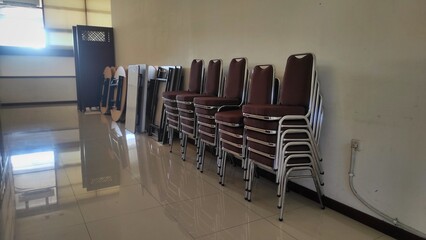 Stacks of brown cushioned chairs and folded tables are neatly arranged against a clean wall in a well-lit, glossy-tiled room, ready for future use.