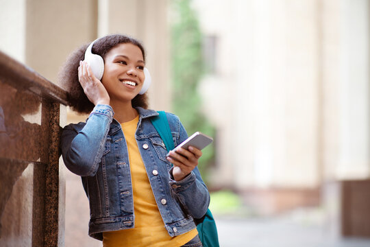Cheerful black female student listening to music with headphones outdoors, smiling broadly, enjoying relaxed moment holding smartphone, casually leaning against a wall on campus - Powered by Adobe